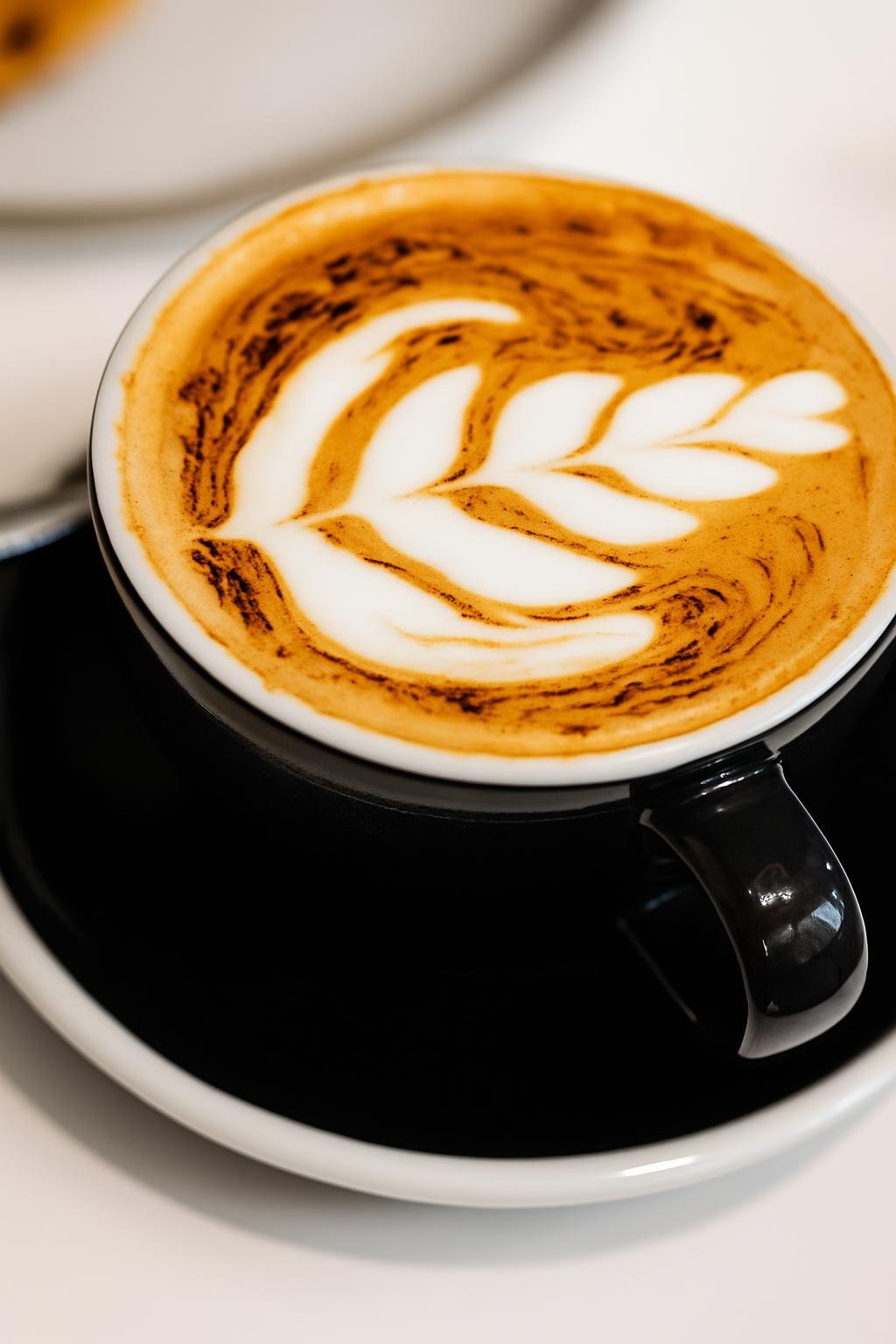 A cappuccino with rosetta latte art in a black cup and saucer on a white table at First Chapter